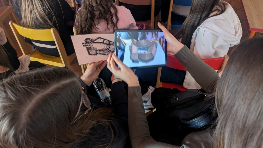 Na fotografiji sta dve osnovnošolki. Ena v rokah pred seboj drži skico zgradbe na listu papirja, druga pa virtualen model iste zgradbe na pametni napravi.