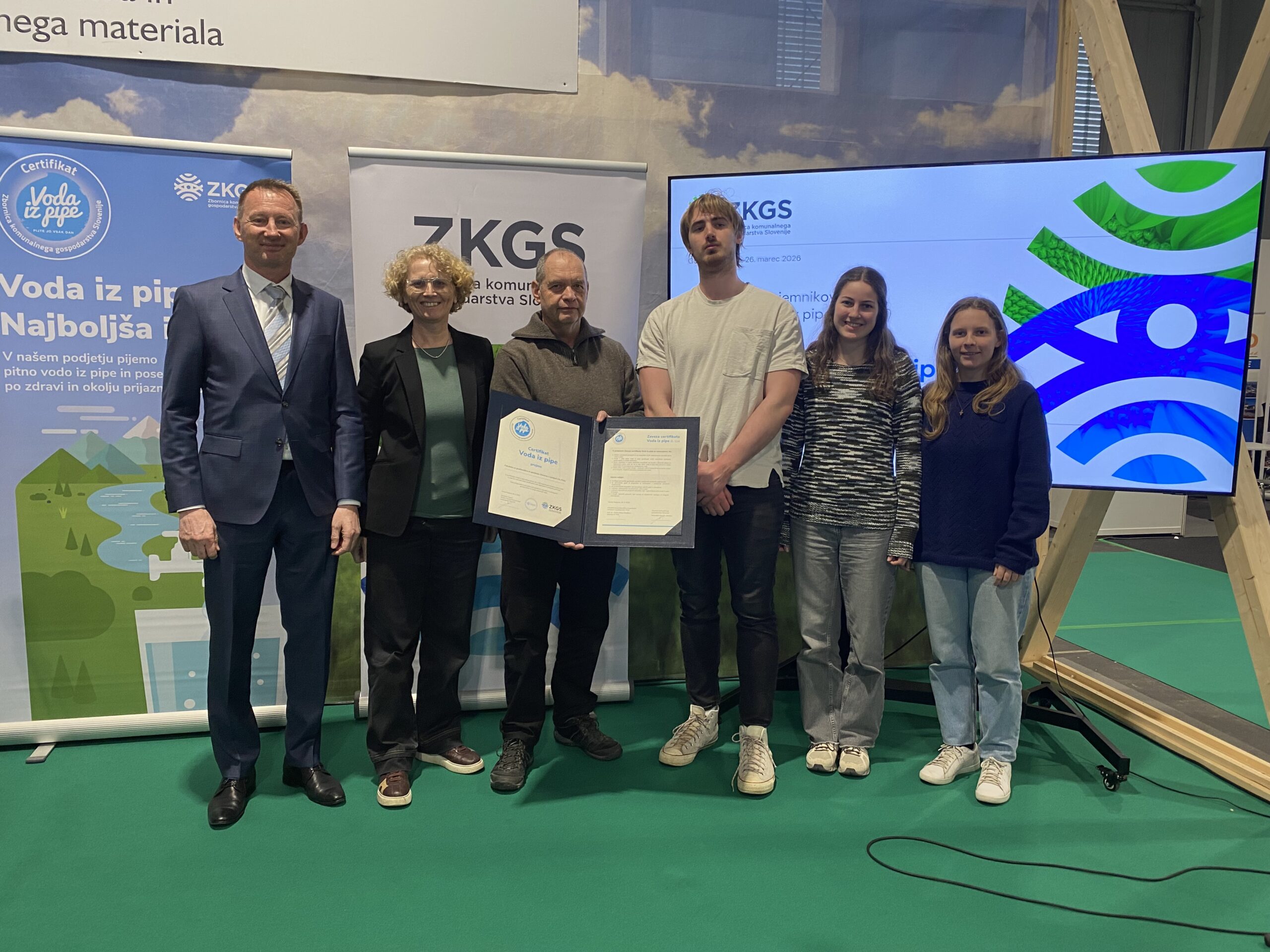 Šest oseb pred zaslonom in pingvinom ZKGS, eden v roki drži certifikat