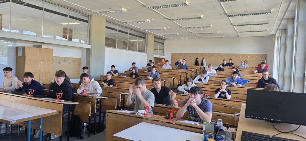 Pogled s katedra na predavalnico. V klopeh sedijo dijaki oziroma tekmovalci, ki rešujejo probleme na področju gradbene mehanike.