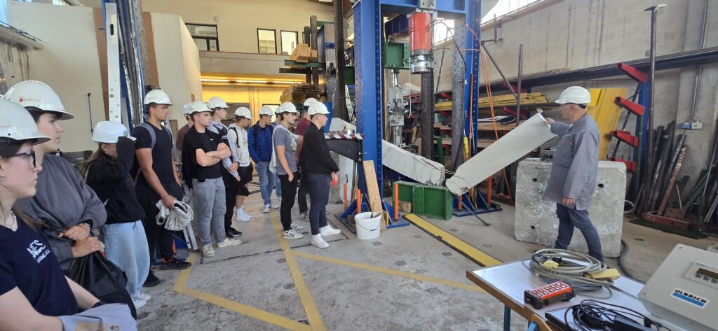 Skupina dijakov stoji v Konstrukcijsko prometnem laboratoriju. Na glavah nosijo čelade. Predavatelj jim razlaga in prikazuje, kako so opravili preizkus betonskega nosilca. Na sliki je viden porušen betonski nosilec.
