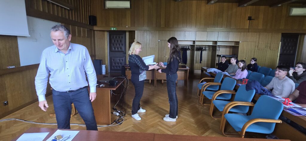 V svečani dvorani UL FGG dekanja čestita najuspešnejši tekmovalki tretjih letnikov. Na stolih v dvorani sedijo ostali tekmovalci, na fotografiji vidimo tudi profesorja, ki dekanji pomaga pri podeljevanju nagrad.