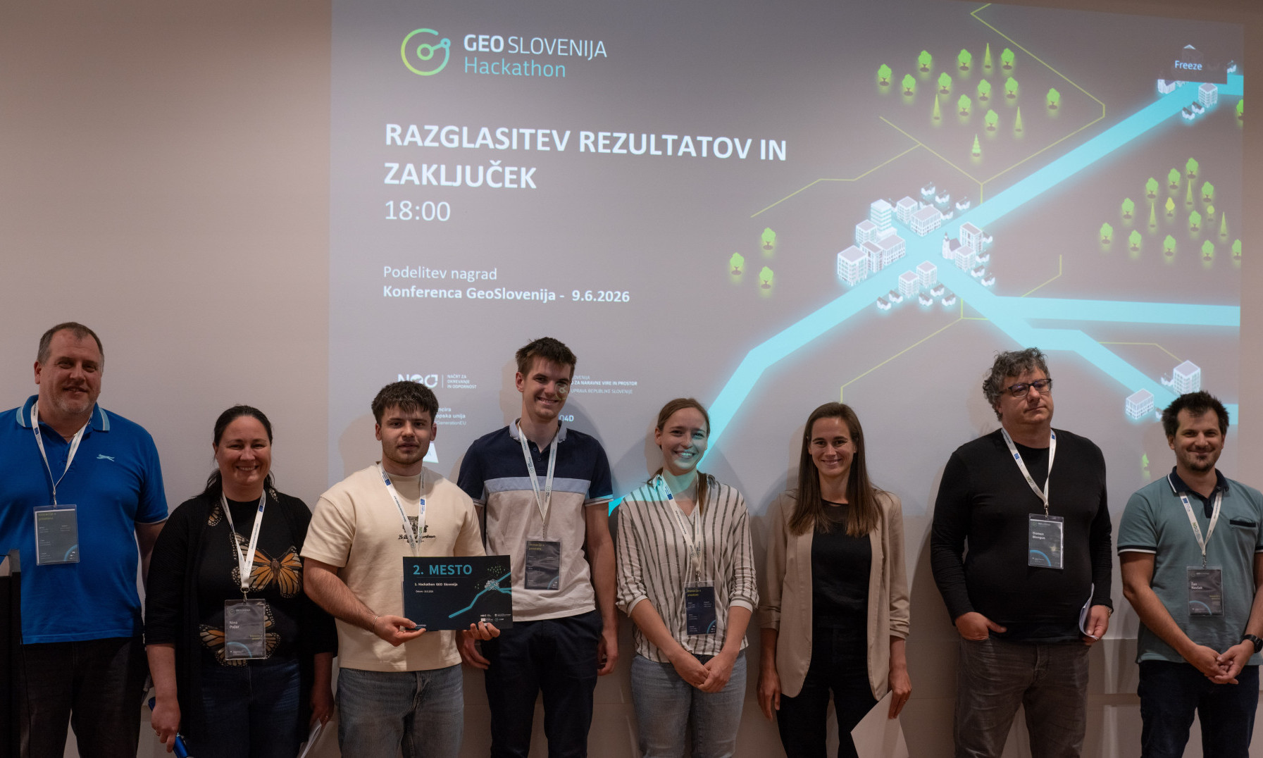 Organizatorji dogodka in študenti UL FGG ob razglasitvi rezultatov Hackathon GEO Slovenija stojijo pred tablo v predavalnici. Eden izmed študentov v rokah drži priznanje za osvojeno 2. mesto.