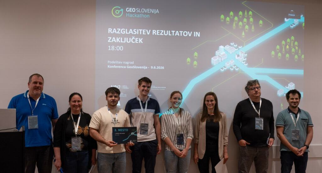 Organizatorji dogodka in študenti UL FGG ob razglasitvi rezultatov Hackathon GEO Slovenija stojijo pred tablo v predavalnici. Eden izmed študentov v rokah drži priznanje za osvojeno 2. mesto.