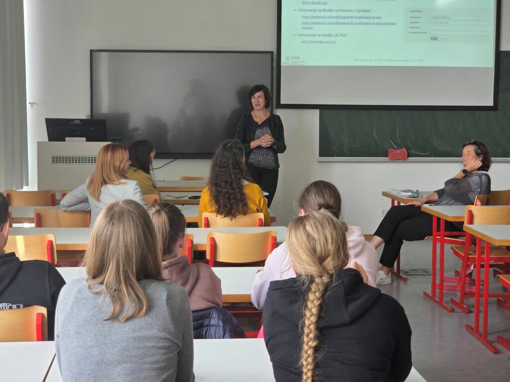 Na fotografiji je pogled na predavalnico iz klopi. Pred tablo je profesorica, ki predavajo, v klopeh pa sedijo obiskovalci informativnega dneva, ki jo poslušajo.