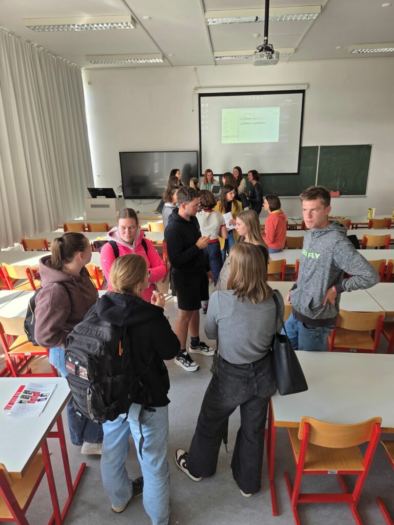 Na fotografiji je pogled na predavalnico z zadnjega dela prostora. Pred tablo vidimo profesorice, pred njimi pa po koncu predavanja stojijo obiskovalci informativnega dneva, ki imajo zanje dodatna vprašanja. Skupina oseb pred fotografom stoji v krogu in se pogovarja.