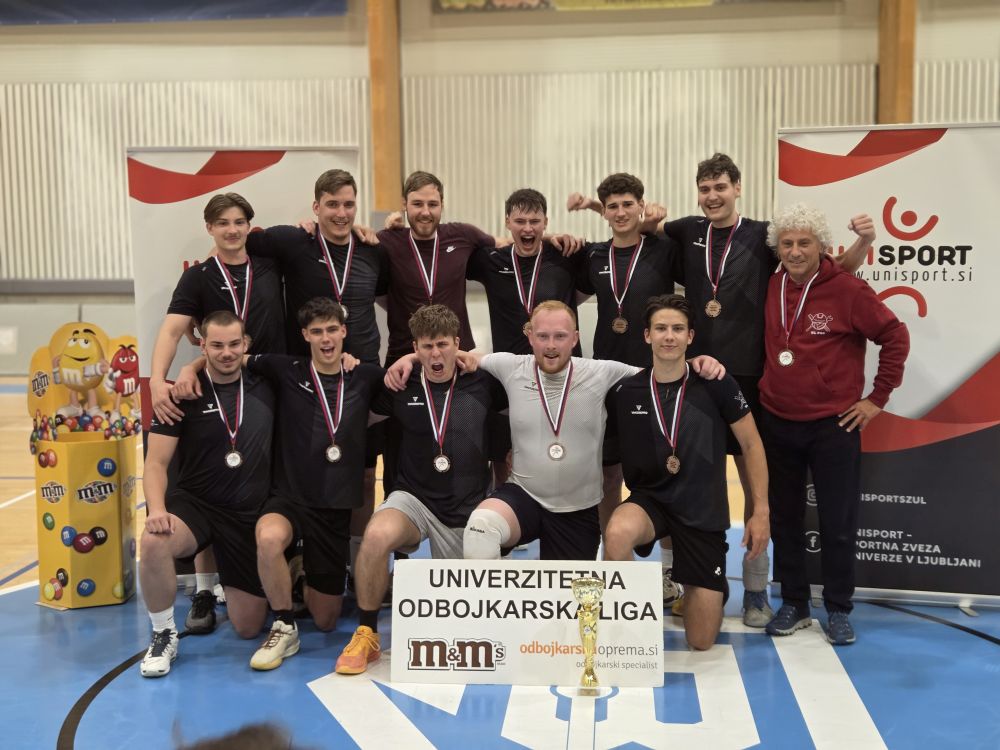 Ekipa 12-ih fantov v dvorani pozira z medaljami okoli vratu. Za njimi sta na vsaki strani pingvina Unisport, zraven njih m&m figura, pred njimi pa pokal ter tabla z napisom Univerzitetna odbojkarska liga in logotipi dveh sponzorjev.