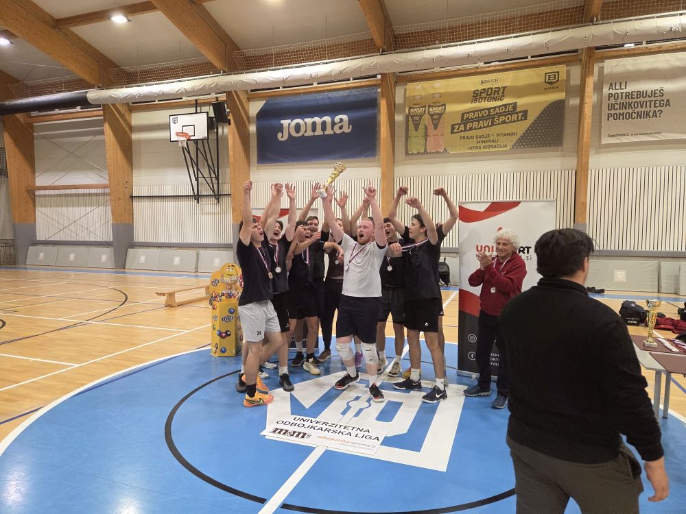 Ekipa FGG se veseli zmage. Fantje so zbrani na kupu, slavijo z vzkliki in rokami v zraku. Okoli vratu imajo medaljo.
