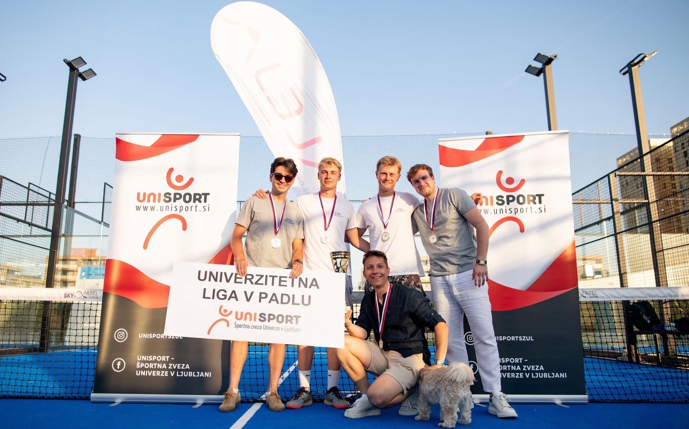 Skupinska fotografija padel ekipe FGG na igrišču. Pet študentov okoli vratu nosi medaljo, eden izmed njih v rokah drži transparent z napisom »Univerzitetna liga v padlu« in logotipom organizatorja. Na fotografiji ob študentih je tudi svetlo rjav pes.v padlu (foto_Športna zveza UL)