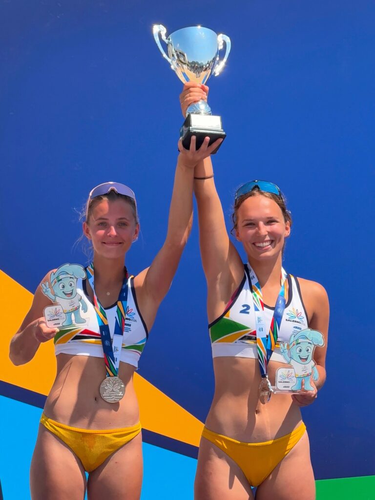Dekleti na zmagovalnem odru držita pokal v zrak, okoli vratu imata medalji, v rokah pa maskoto tekmovanja.