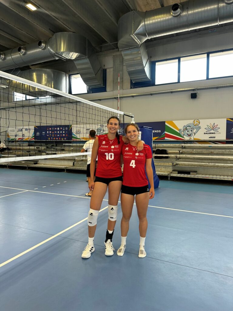 Dekleti pozirata v športni dvorani pred odbojkarsko mrežo.