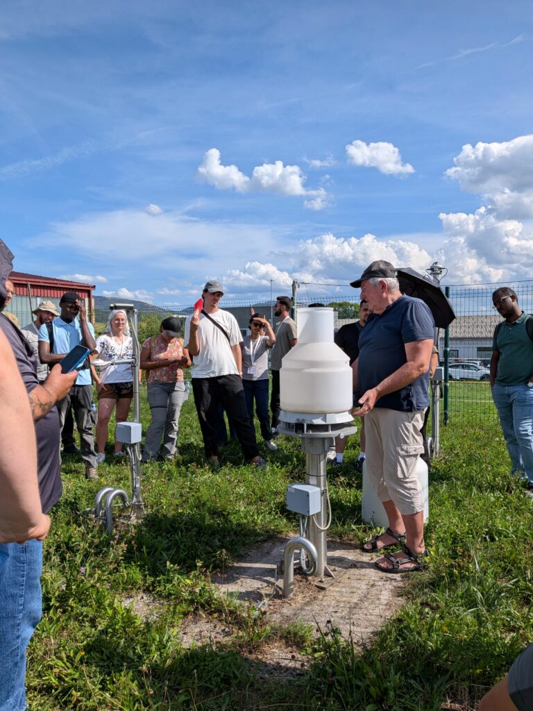 Filip Štucin z Agencije RS za okolje udeležencem poletne šole HydRoData 2026 na avtomatski meteorološki postaji v Postojni predstavlja delovanje pluviografa – kontinuiranega merilnika za spremljanje količine padavin.